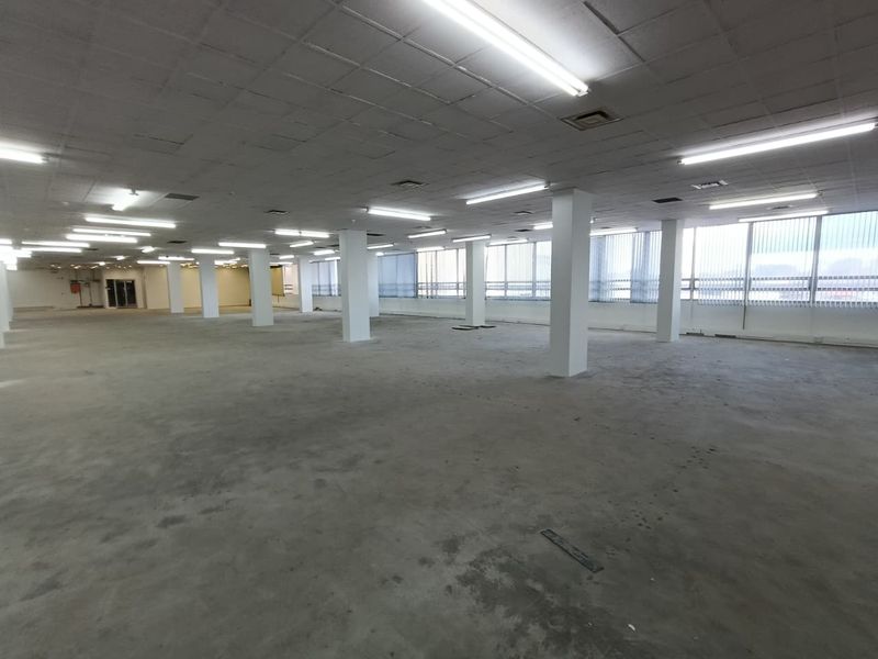 A large, open-plan office space featuring structural columns, suspended ceilings with fluorescent lighting, and expansive windows with vertical blinds.