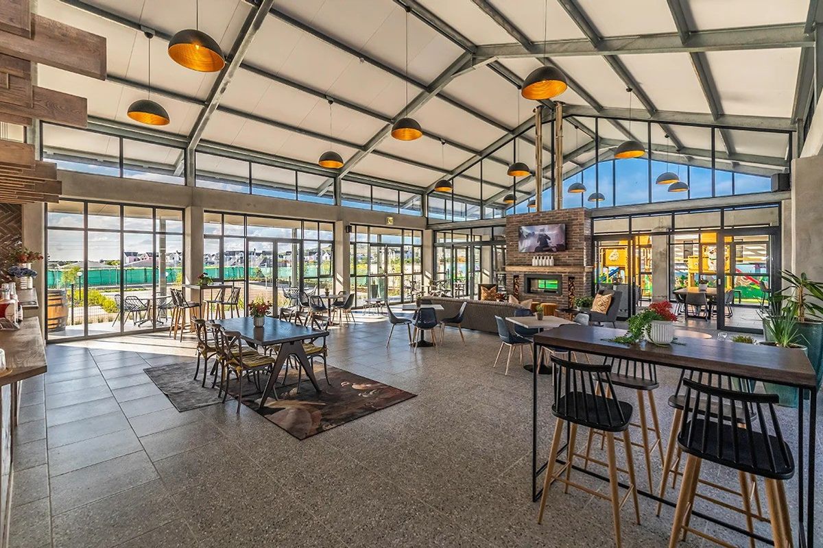 Spacious modern clubhouse featuring high vaulted ceilings with exposed steel trusses, extensive glass walls, tiled flooring, and a central brick fireplace.