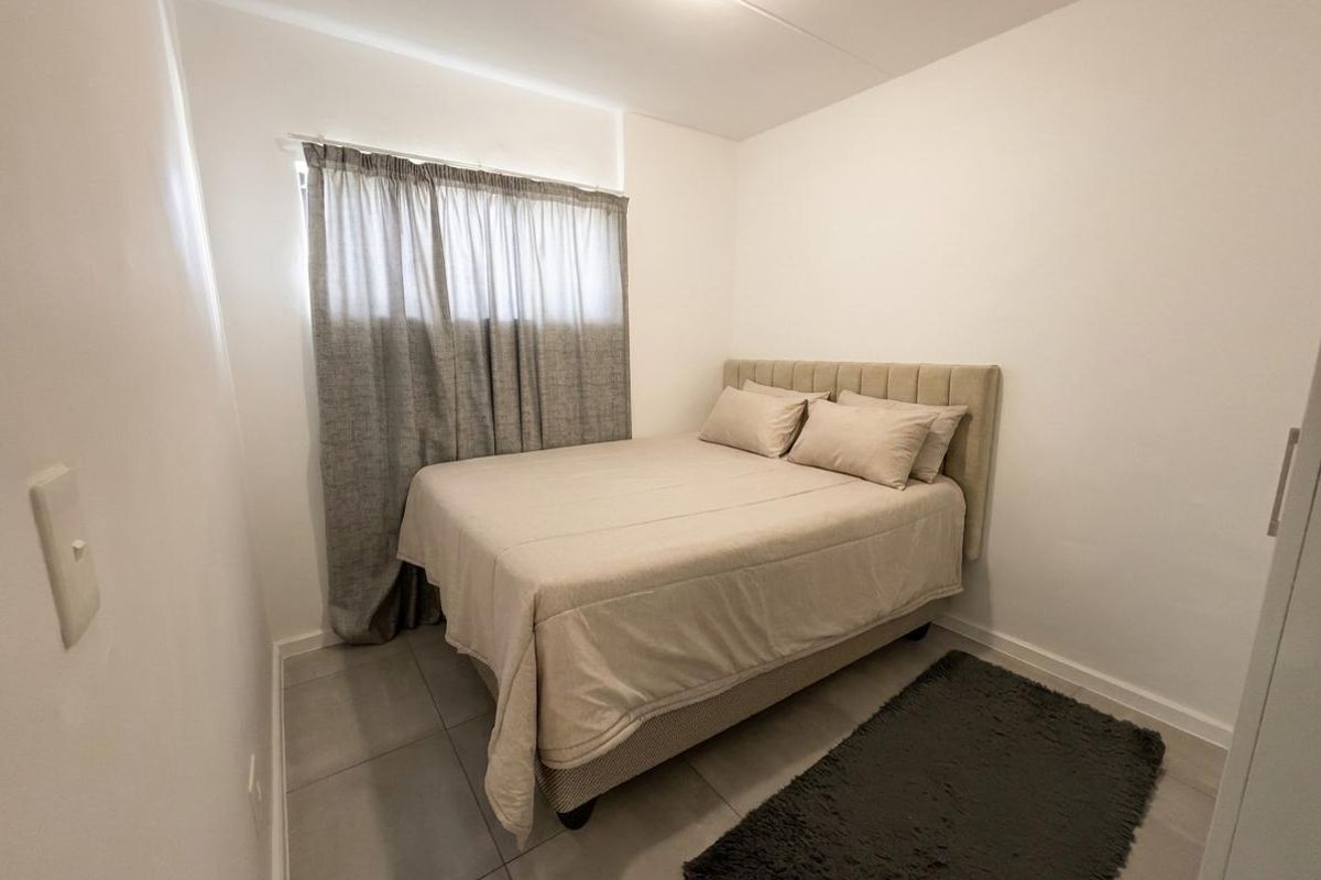A compact bedroom featuring neutral tiled flooring, white walls, and a window with fitted curtain tracks.