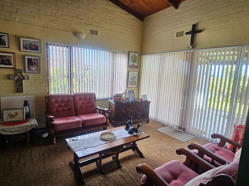 A living room featuring exposed timber ceilings, painted brick walls, and large windows with vertical blinds, offering a dated but functional space.