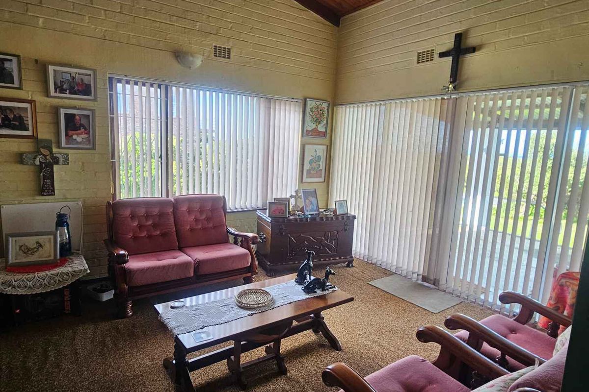 A living room featuring exposed timber ceilings, painted brick walls, and large windows with vertical blinds, offering a dated but functional space.