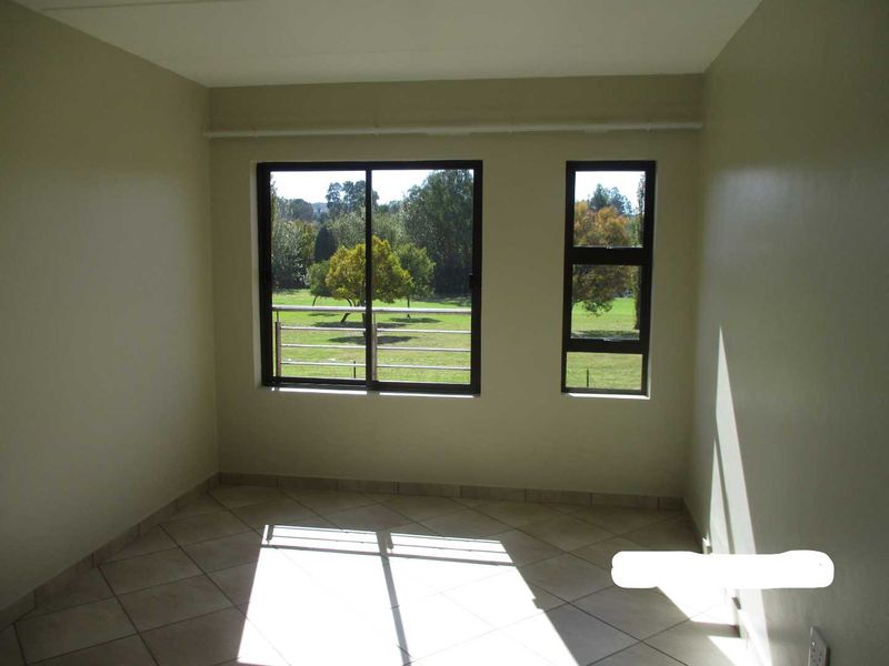 A bright, empty bedroom featuring neutral wall tones, tiled flooring, and large windows offering a pleasant green garden view.