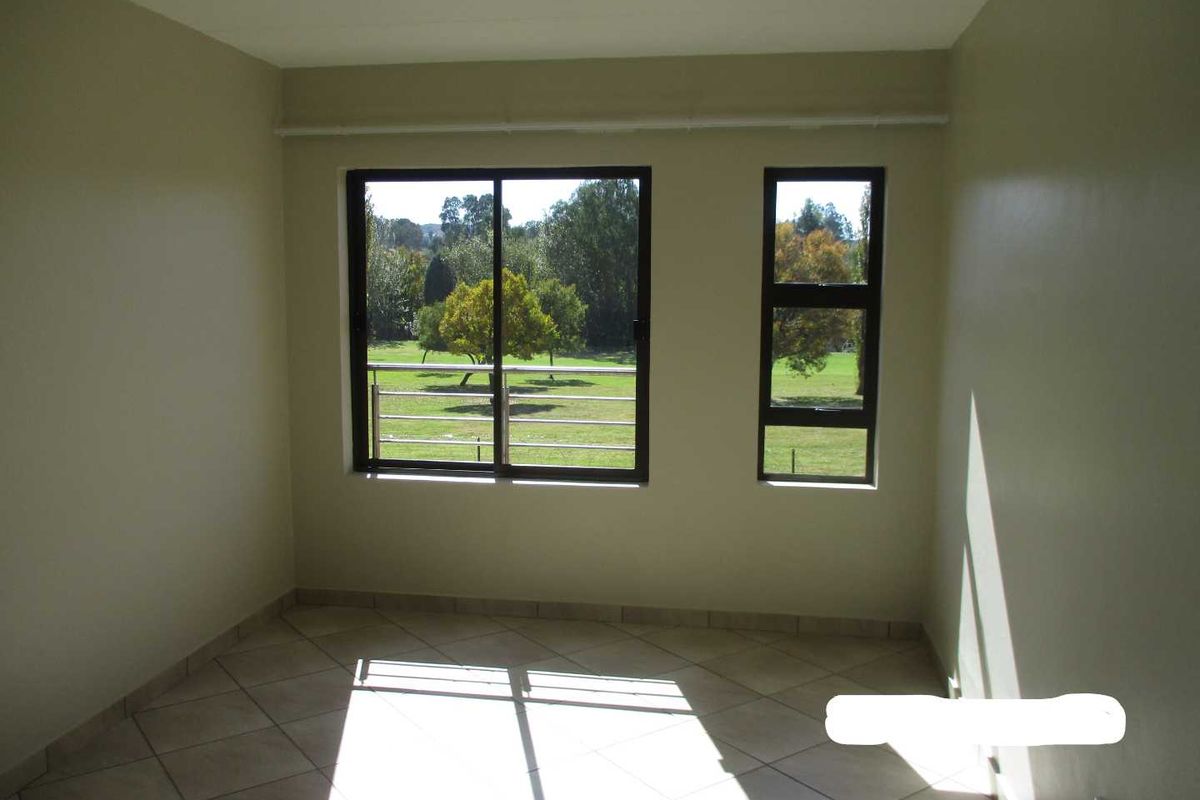 A bright, empty bedroom featuring neutral wall tones, tiled flooring, and large windows offering a pleasant green garden view.