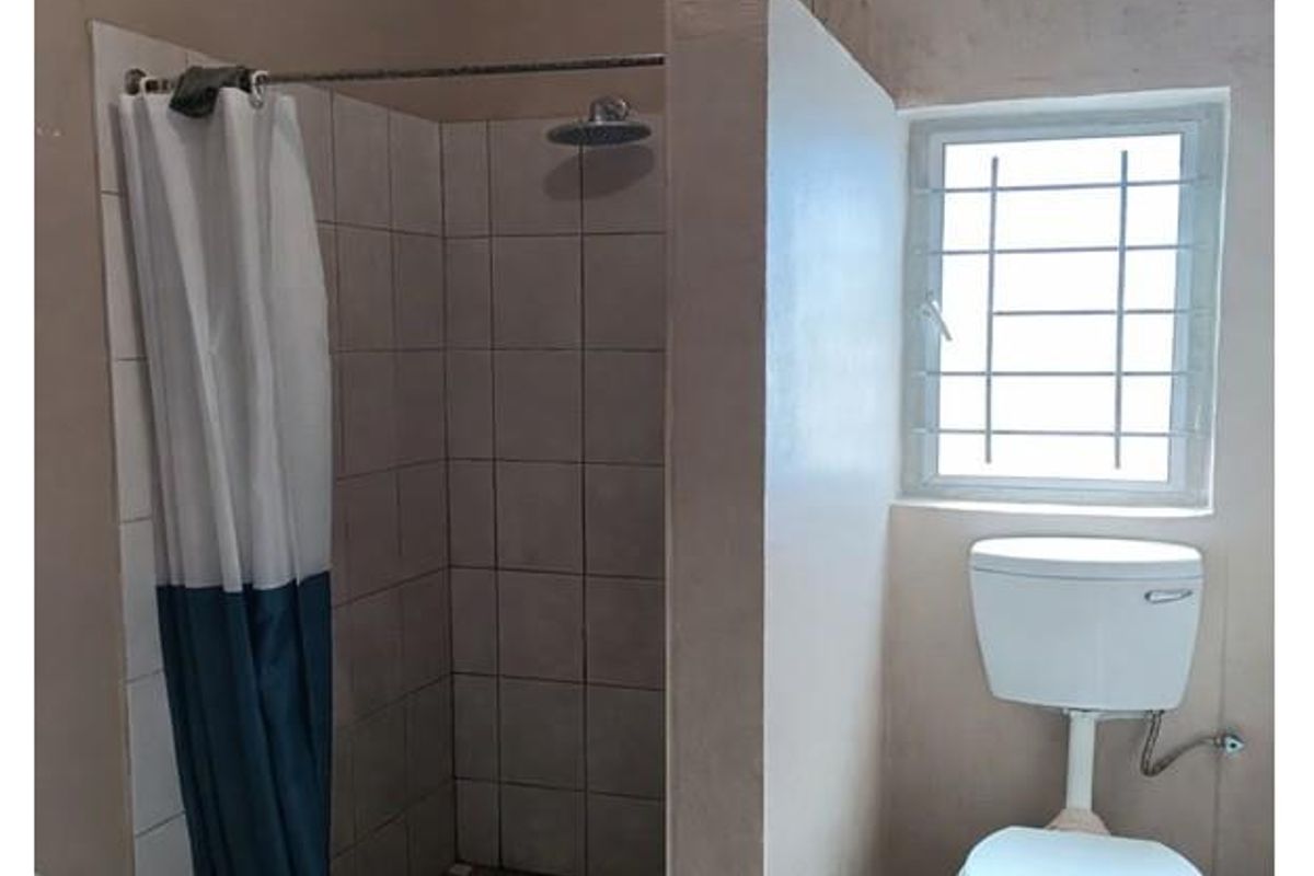 A functional bathroom featuring a walk-in shower with tiled surround and a standard toilet, finished with neutral wall tones and a security-grilled window.