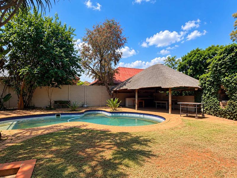 Outdoor pool area featuring a thatched lapa with a built-in braai, surrounded by a garden and boundary wall.