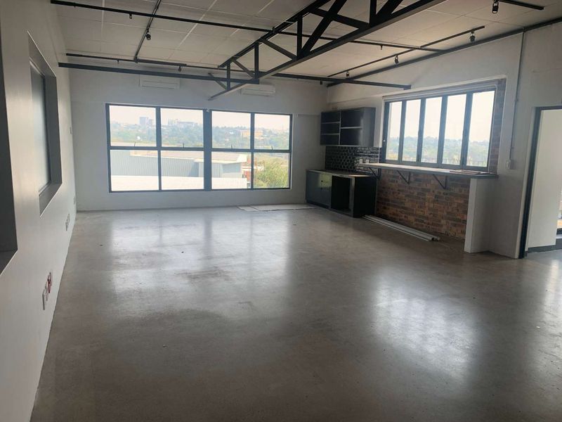 Spacious open-plan office area featuring polished concrete floors, exposed industrial ceiling trusses, large windows with views, and a built-in kitchenette area.