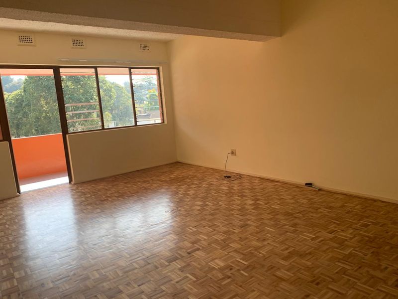 A spacious, empty living room featuring polished parquet flooring, a large sliding glass door leading to a balcony, and a structural ceiling beam.