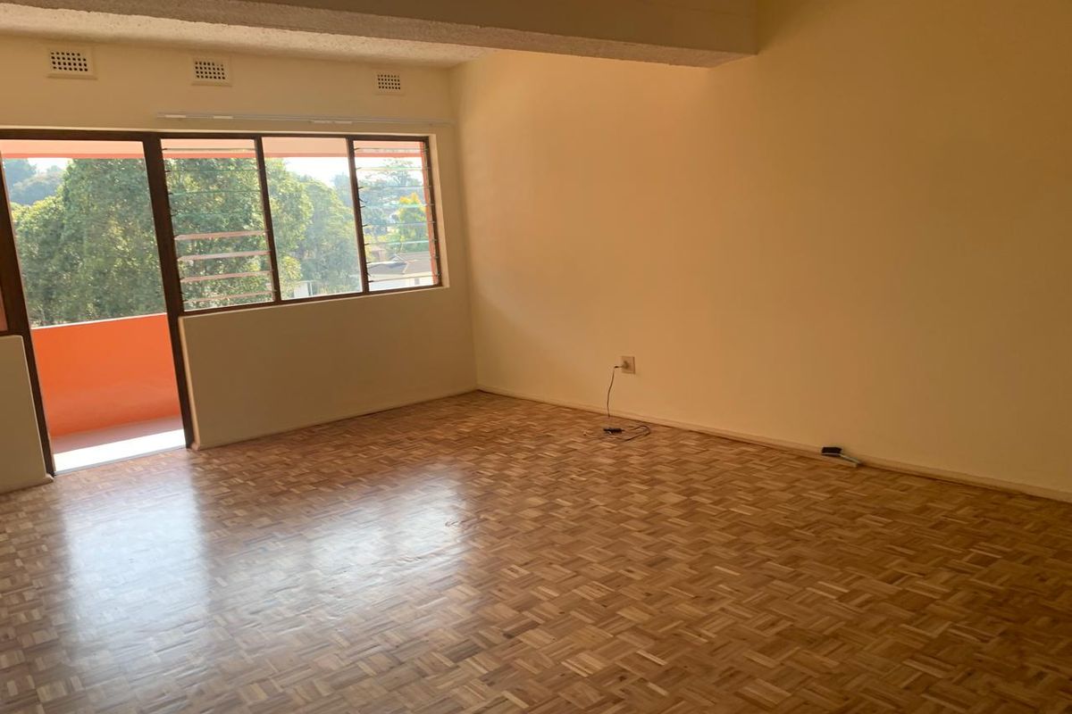 A spacious, empty living room featuring polished parquet flooring, a large sliding glass door leading to a balcony, and a structural ceiling beam.