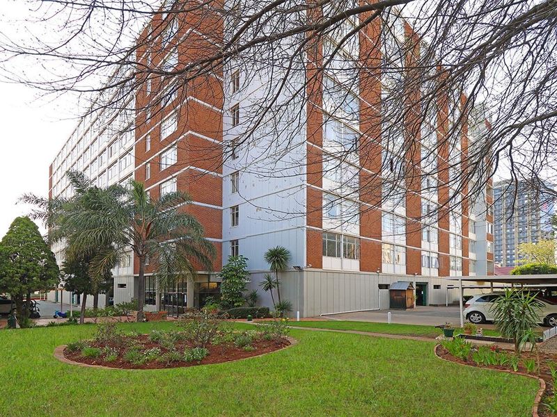 Exterior view of a multi-story apartment building featuring a brick and panel facade, surrounded by a landscaped garden and covered parking area.