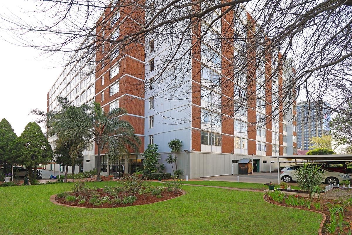 Exterior view of a multi-story apartment building featuring a brick and panel facade, surrounded by a landscaped garden and covered parking area.
