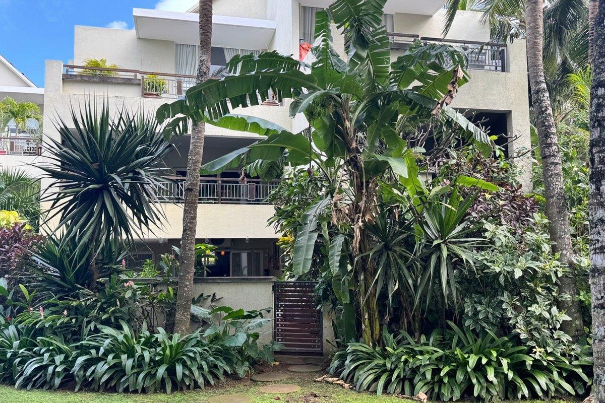 Rear view of a multi-story apartment building featuring private balconies, lush tropical landscaping, and a garden pathway.