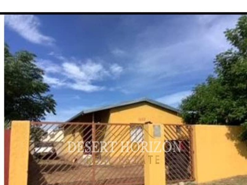 Front view of a single-story house featuring a yellow facade, pitched roof, and a gated perimeter wall.
