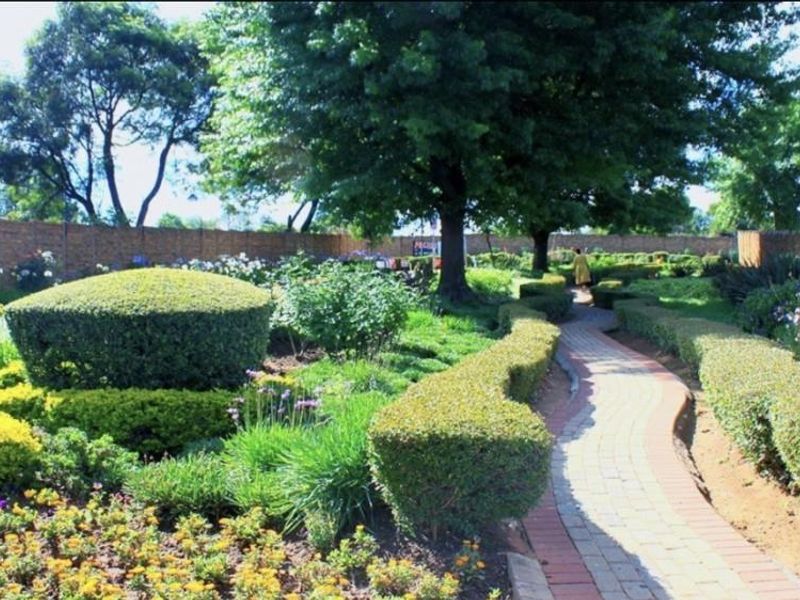 A well-maintained garden featuring a paved walkway, manicured hedges, mature trees, and a brick boundary wall.