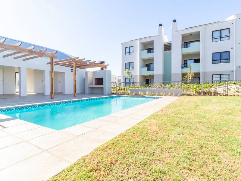 Communal pool area featuring a paved deck, timber pergola, and built-in braai facility, set within a well-maintained apartment complex garden.