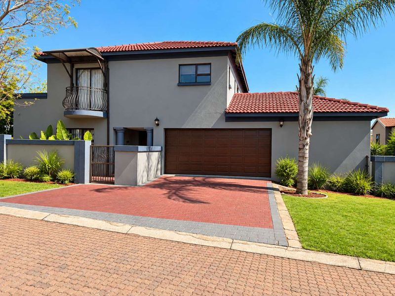 A well-maintained double-story home featuring a paved driveway, double garage, and a balcony with wrought iron detailing, set within a neat landscaped garden.