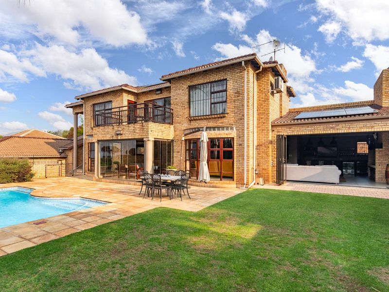 Rear view of a double-story brick house featuring a swimming pool, landscaped lawn, tiled patio area, and an upper-level balcony.
