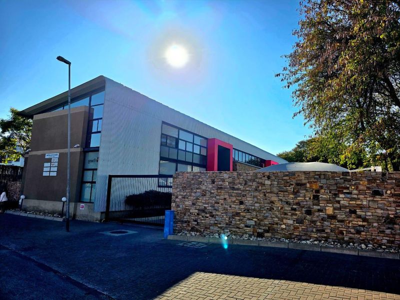 Front exterior view of a modern office building featuring a stone boundary wall, metal security gate, and corrugated metal facade.