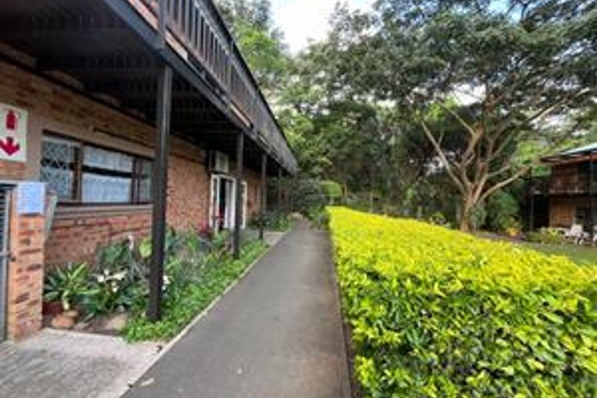 A paved walkway leads along the exterior of a brick retirement village building, featuring a covered veranda and landscaped garden surroundings.