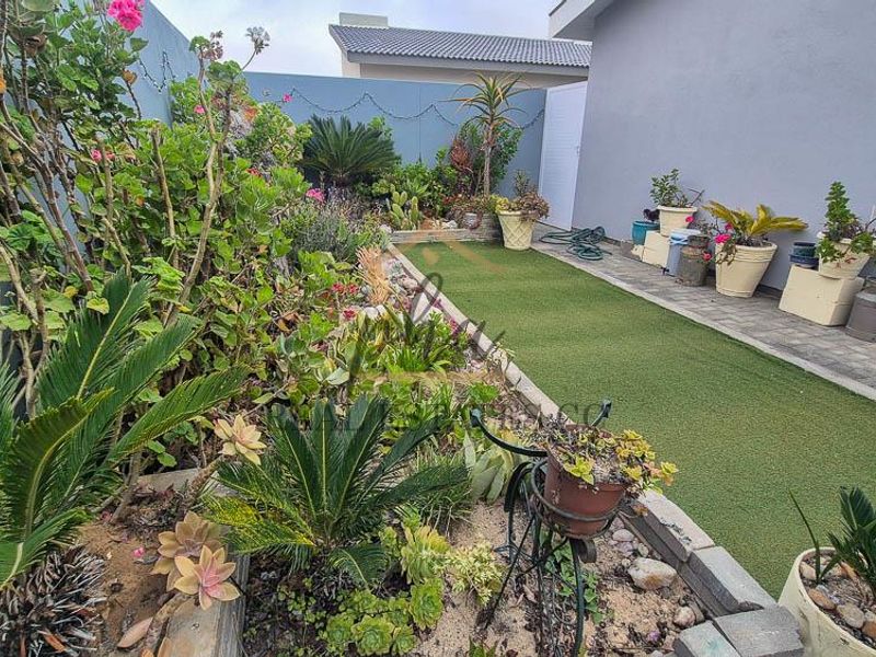 A well-maintained side garden featuring artificial turf, a paved walkway, and established garden beds enclosed by a boundary wall.