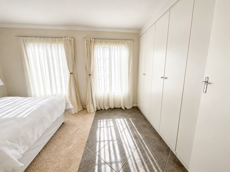 A bright bedroom featuring neutral walls, tiled flooring, and extensive built-in cupboard storage with natural light filtering through dual windows.