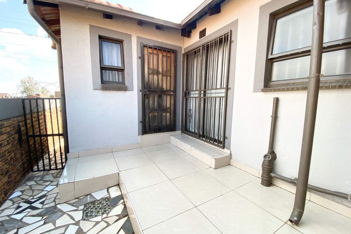 A paved rear patio area featuring tiled flooring, metal security gates on doors, and exposed exterior drainage infrastructure.