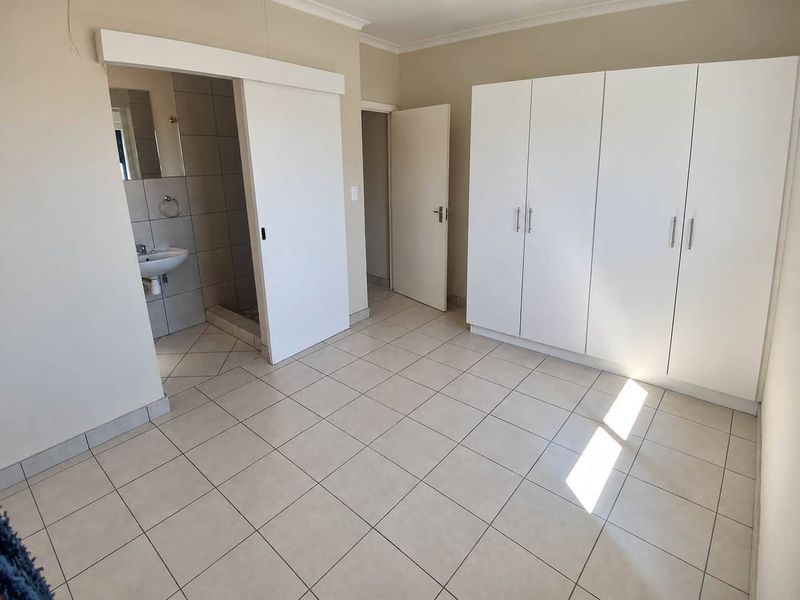A spacious bedroom featuring neutral tiled flooring, built-in white cupboards, and an adjoining en-suite bathroom.