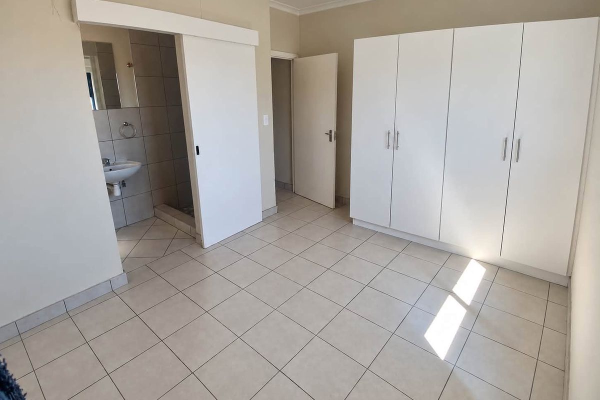 A spacious bedroom featuring neutral tiled flooring, built-in white cupboards, and an adjoining en-suite bathroom.