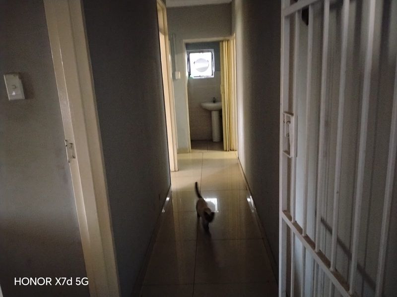 A narrow hallway with tiled flooring leading towards a bathroom area featuring a pedestal basin and a security gate.