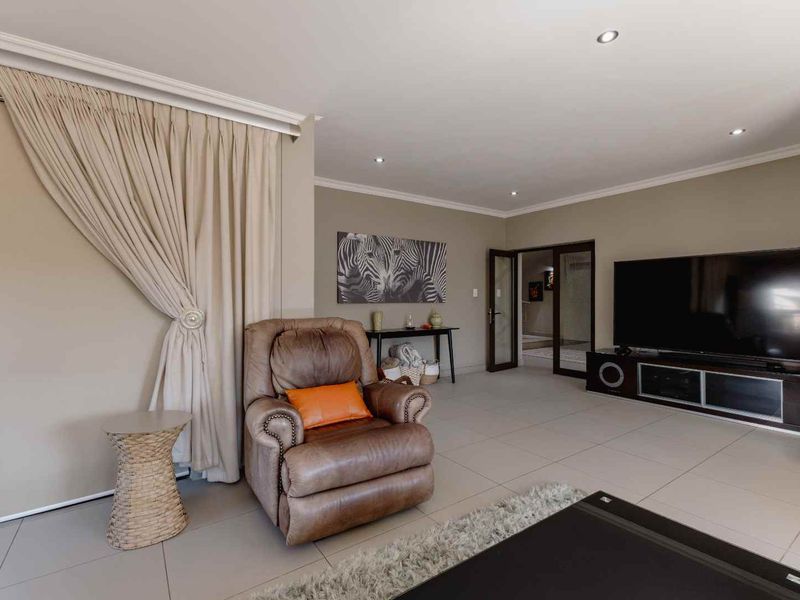 A spacious living room featuring neutral tiled flooring, recessed downlighting, and glass panelled doors leading to adjacent areas.