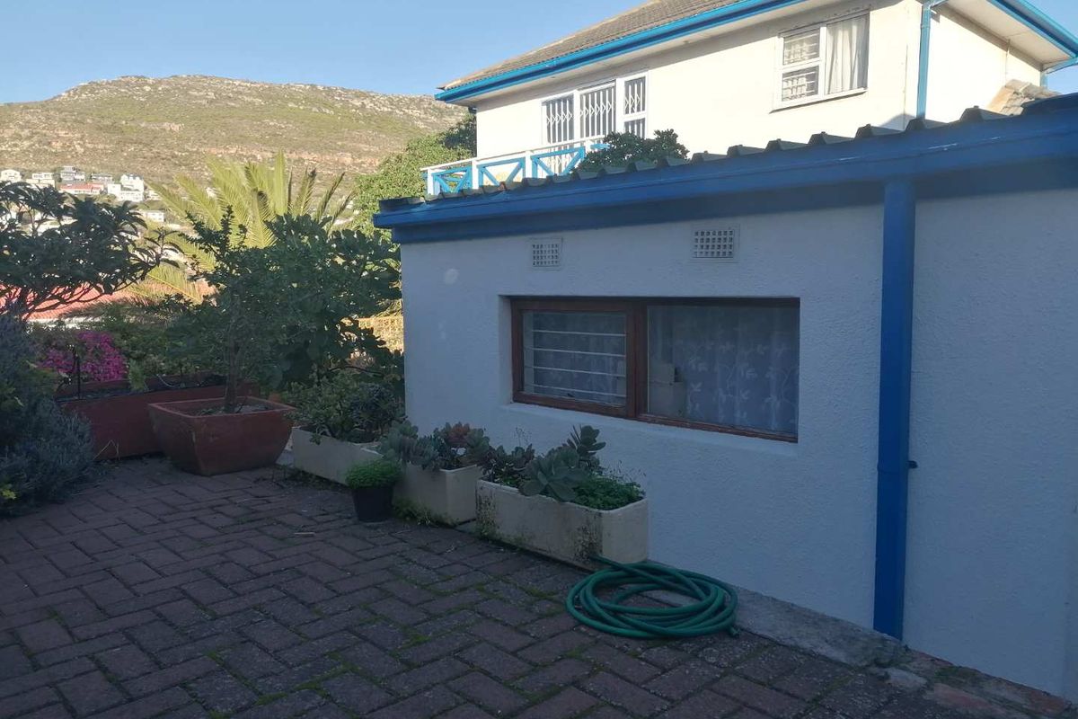 A view of the cottage exterior featuring a paved patio area, white rendered walls, and blue trim, set against a mountain backdrop.