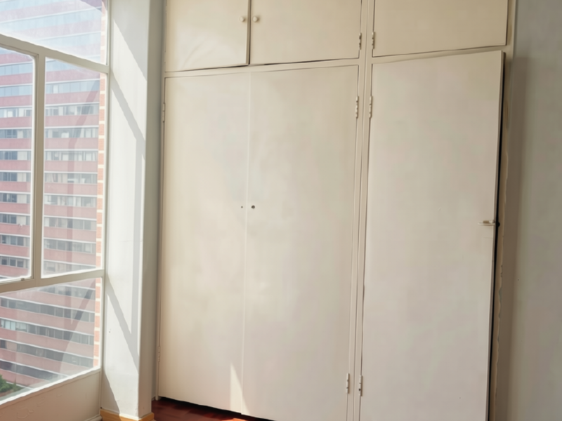 A bright, empty bedroom featuring built-in cupboards, timber flooring, and a large window with an urban view.