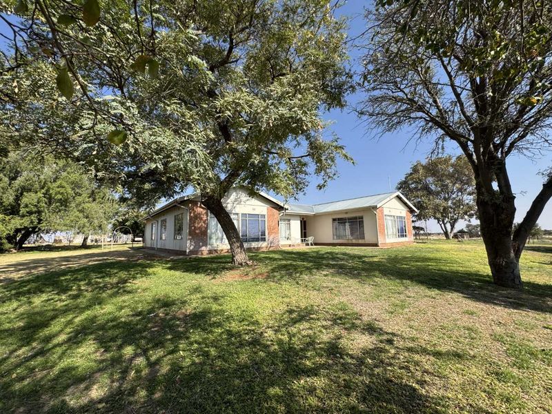 A view of the rear brick farmhouse elevation, featuring large windows, a metal roof, and expansive surrounding lawn with mature trees.