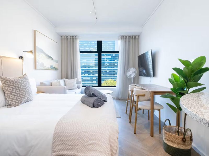 A well-lit bedroom featuring herringbone flooring, a large window with city views, and a wall-mounted television.