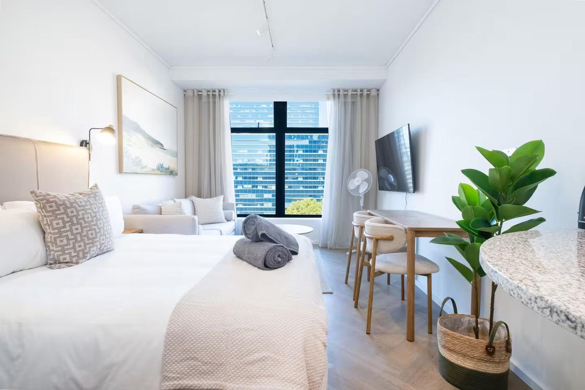 A well-lit bedroom featuring herringbone flooring, a large window with city views, and a wall-mounted television.