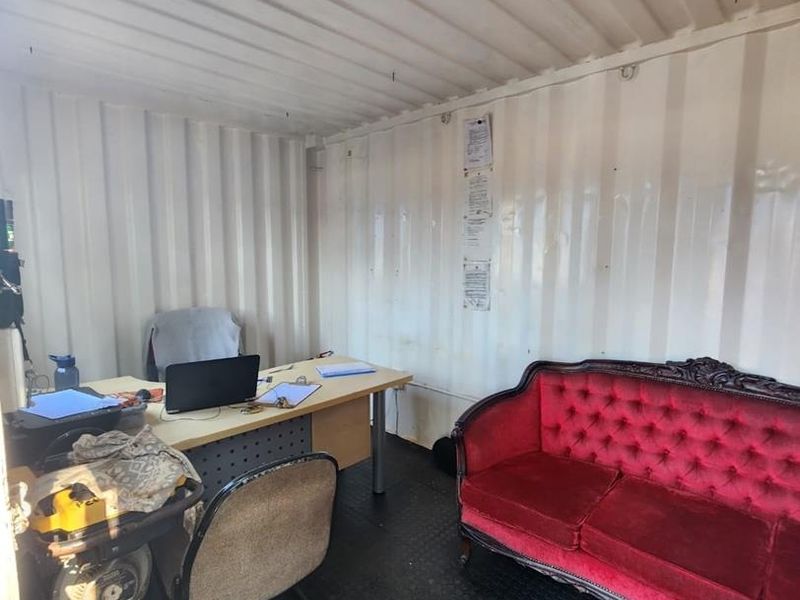 A functional office space housed within a converted shipping container, featuring corrugated metal walls, durable rubber flooring, and overhead fluorescent lighting.