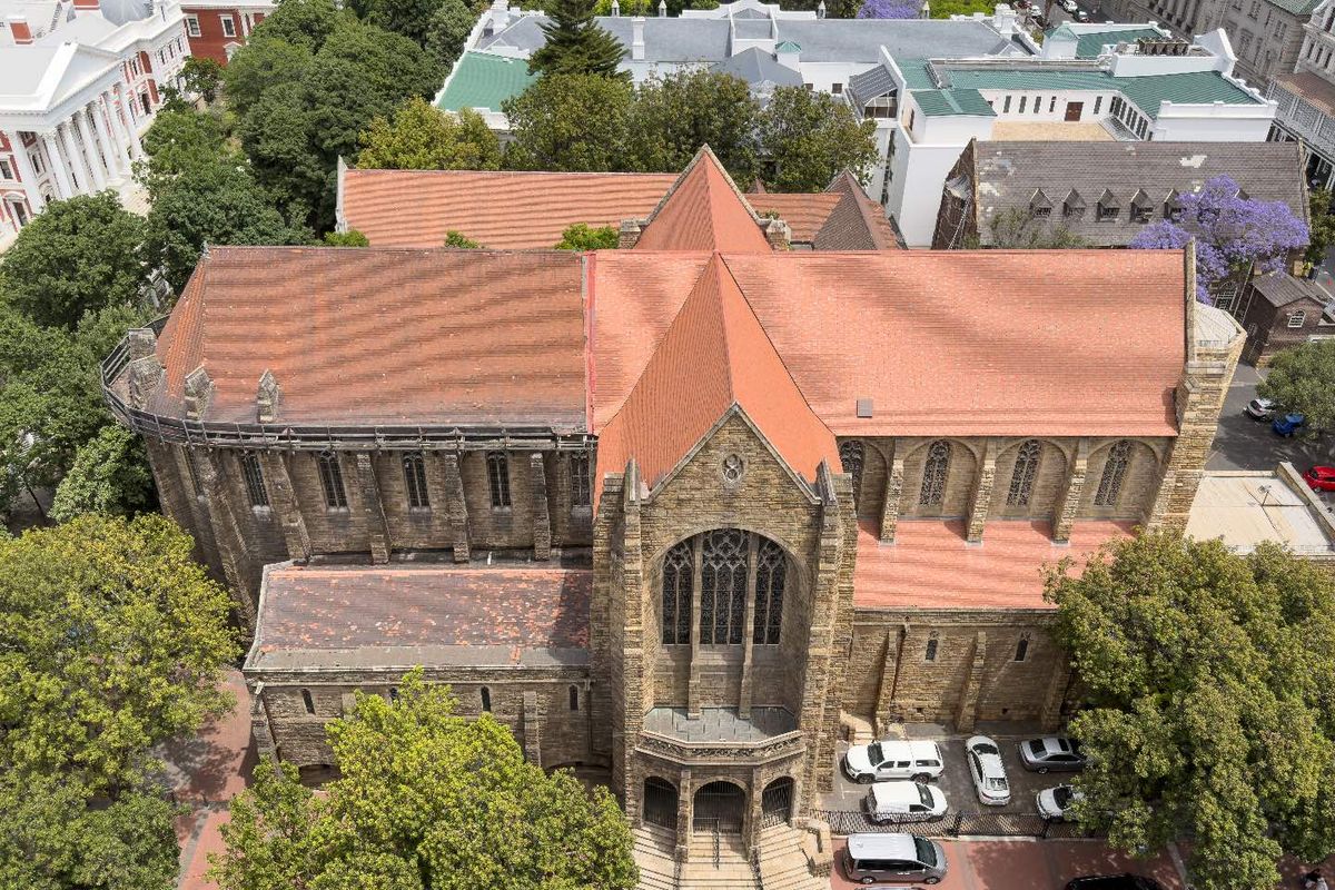 Aerial view of a historic stone building featuring a prominent tiled roof, arched architectural details, and surrounding mature trees in an urban setting.