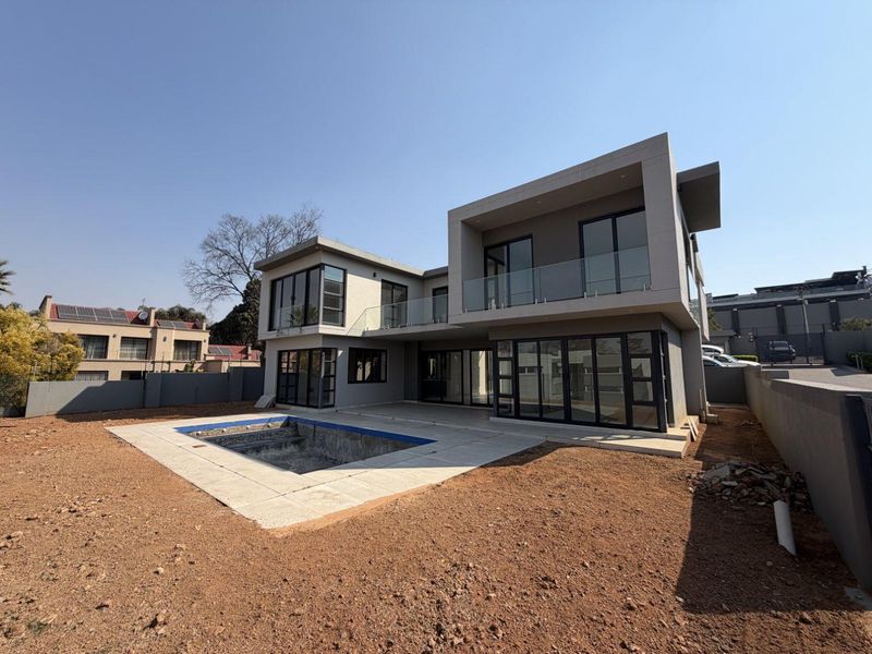 Rear view of a modern double-story house featuring a swimming pool, glass balcony railings, and large sliding doors, set within an unfinished garden area.
