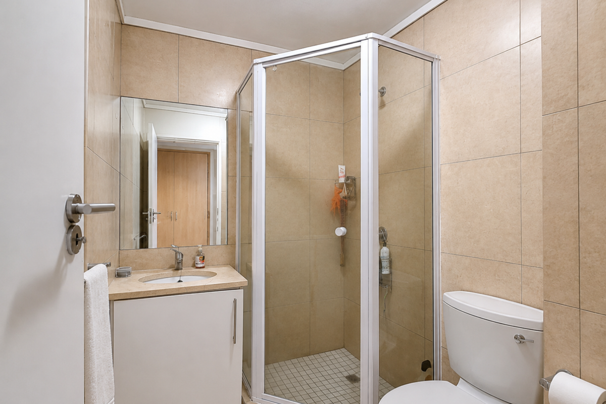 A functional bathroom featuring a corner shower enclosure, a vanity unit with an integrated basin, and neutral wall tiling.