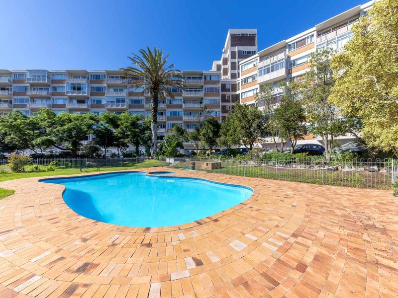 A well-maintained communal swimming pool area featuring brick paving, landscaped gardens, and metal perimeter fencing set against a large apartment building.