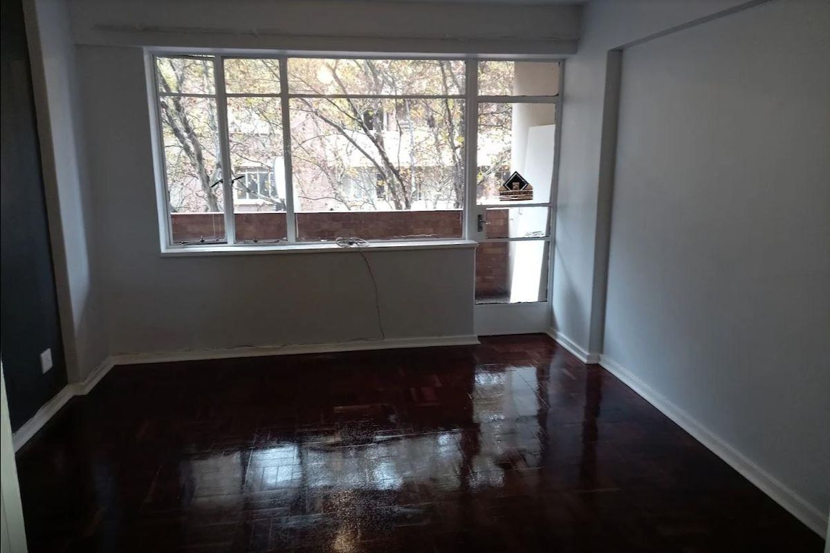 A bright, empty living room featuring polished dark parquet flooring, large windows, and direct access to an outdoor balcony area.