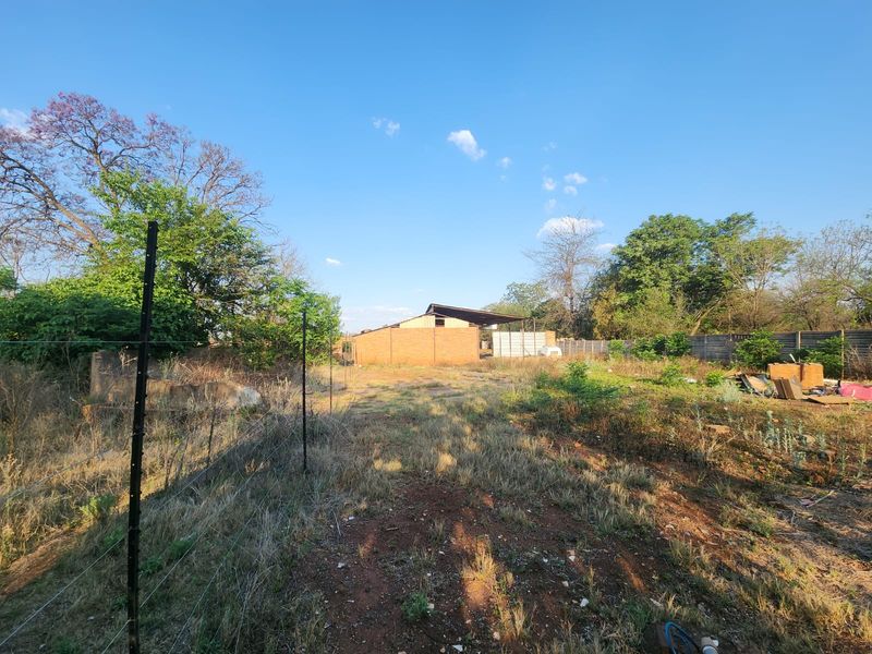 A large, undeveloped outdoor area featuring a wire fence, overgrown vegetation, and a basic brick structure with a metal roof in the background.