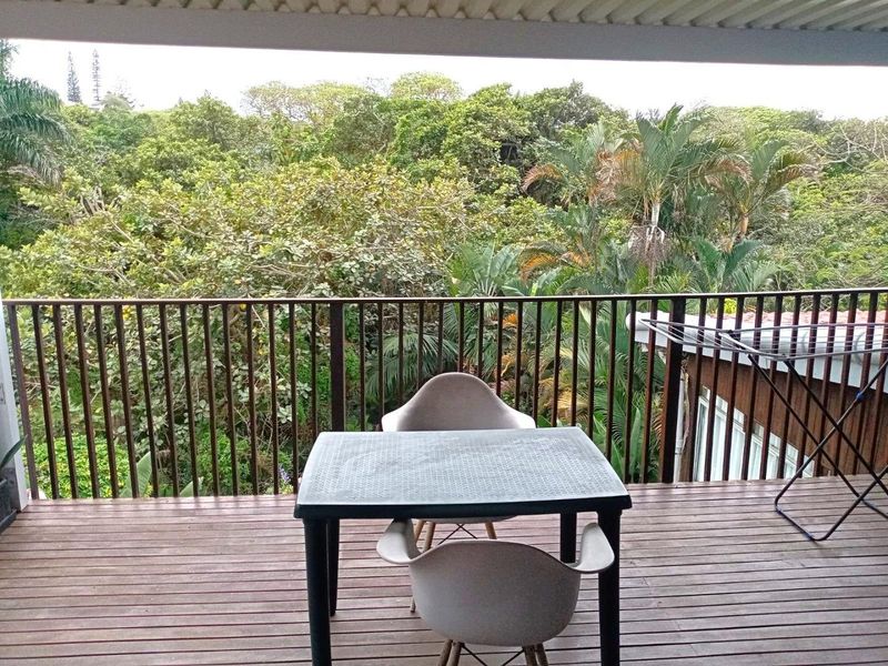 A covered balcony featuring timber decking and a metal balustrade, offering a pleasant elevated view of the surrounding lush greenery.
