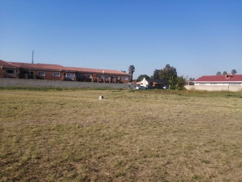 A vacant residential plot of land featuring open grass, a boundary wall, and surrounding residential properties in a quiet neighborhood setting.