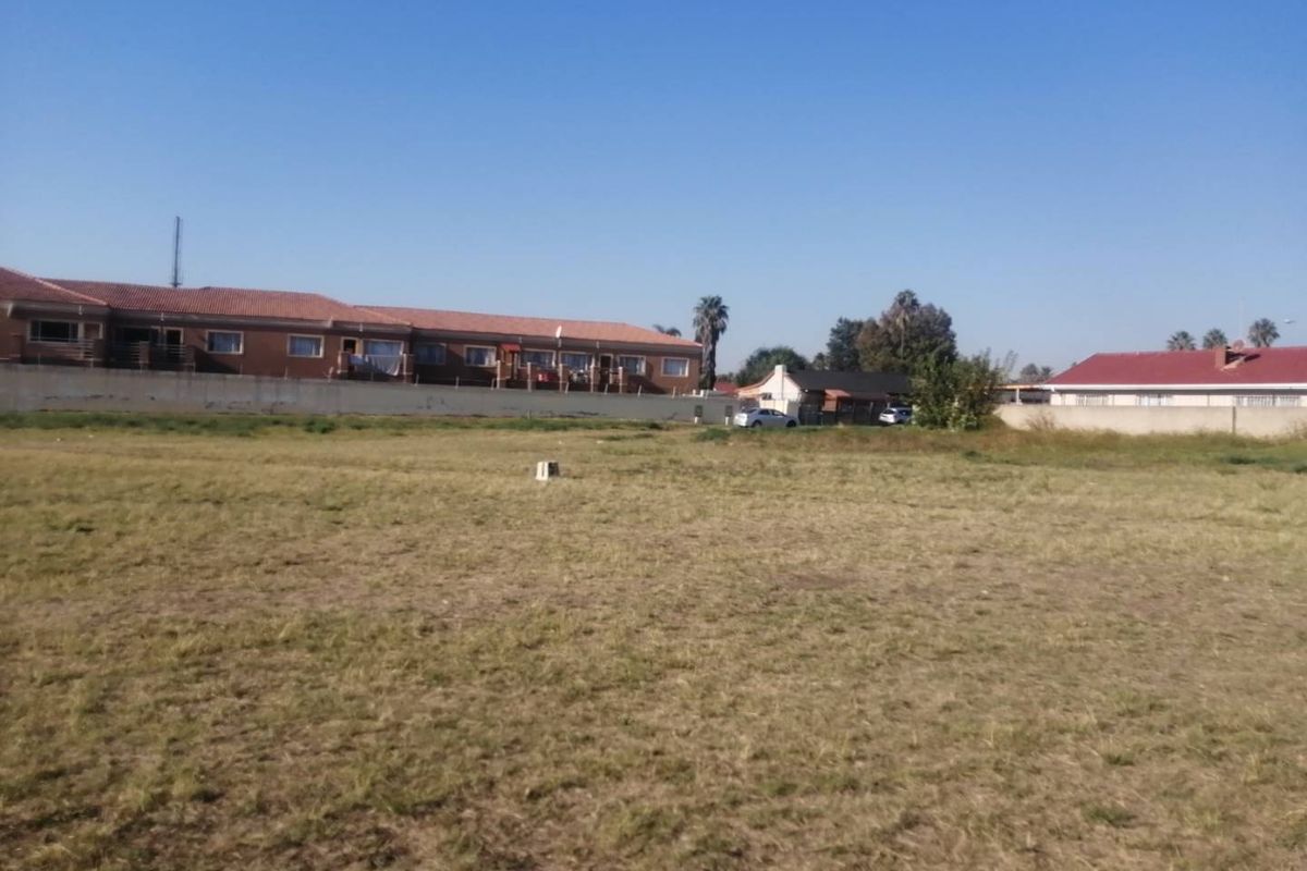 A vacant residential plot of land featuring open grass, a boundary wall, and surrounding residential properties in a quiet neighborhood setting.