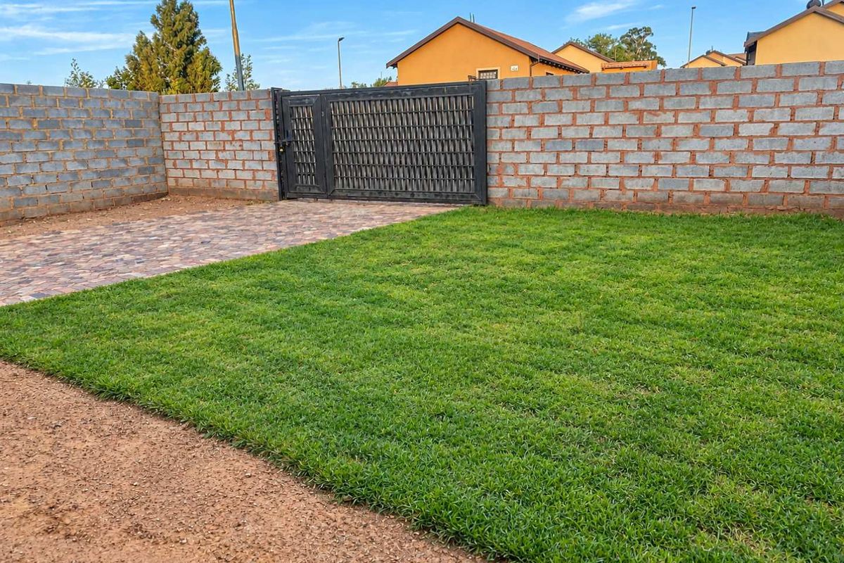 A private rear garden area featuring a well-maintained lawn, paved driveway section, and secure metal gate access enclosed by brick boundary walls.