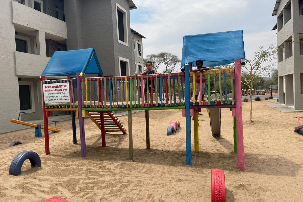 Communal playground area situated between apartment blocks, featuring fixed wooden climbing structures and a sand surface.