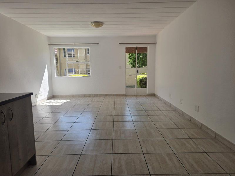A bright, empty open-plan living area featuring neutral tiled flooring, clean white walls, and direct access to an outdoor space through a glass door.
