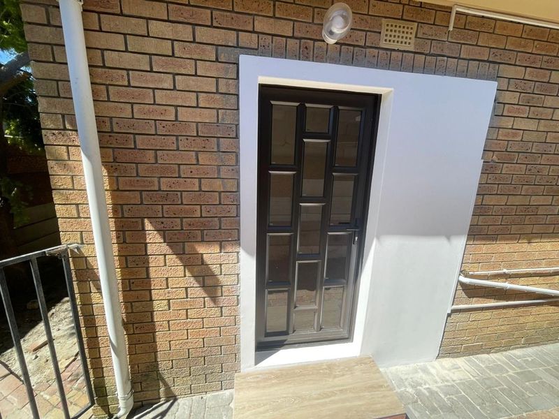 A rear exterior view of the apartment featuring a modern glass panel door set against a brick facade with paved flooring.