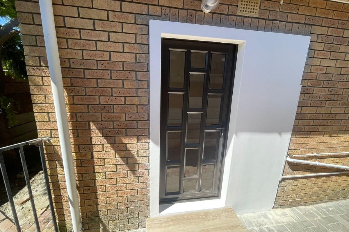 A rear exterior view of the apartment featuring a modern glass panel door set against a brick facade with paved flooring.
