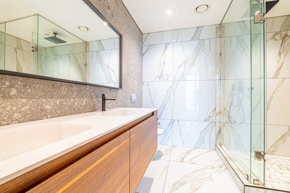 Modern bathroom featuring a double vanity with wood cabinetry, large framed mirror, and a spacious walk-in glass shower with marble-look wall tiles.